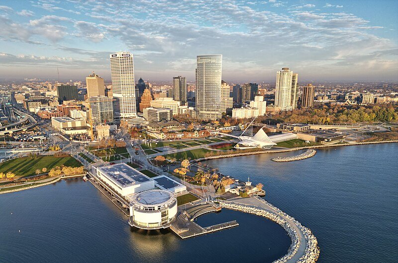 Aerial view of a city skyline at sunset, featuring modern skyscrapers, a waterfront with a white circular building, landscaped parks, and a glowing urban backdrop.