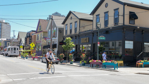 "A vibrant street scene featuring shops, cafes, and restaurants with outdoor seating, colorful flower planters, and a person riding a bicycle across the intersection.