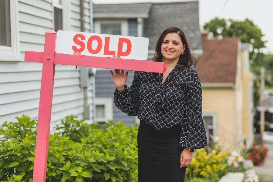Smiling woman standing next to a 'Sold' sign in front of a house, representing the successful sale of a property.