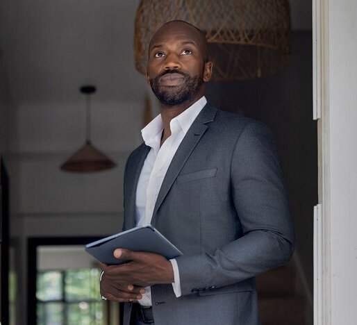 Professional real estate agent in a suit, holding a tablet and standing at the doorway of a stylish home, ready to showcase property details to potential buyers.