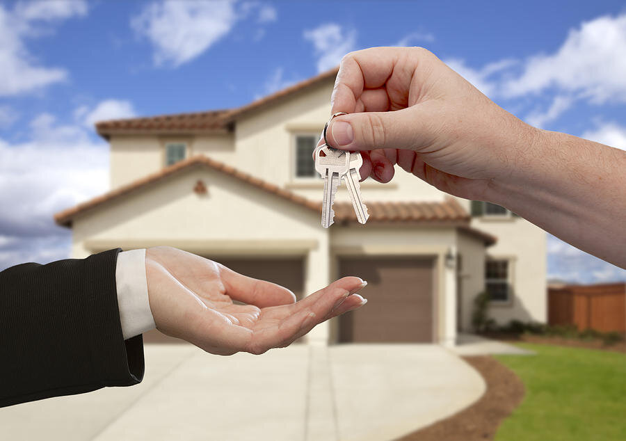 A close-up of a hand passing house keys to another hand in front of a modern, two-story home, symbolizing the finalization of a real estate transaction.