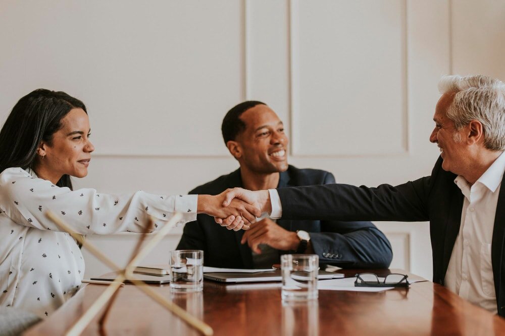 A diverse group of business professionals in a meeting, with two people smiling and shaking hands across a conference table, indicating a successful deal or agreement.