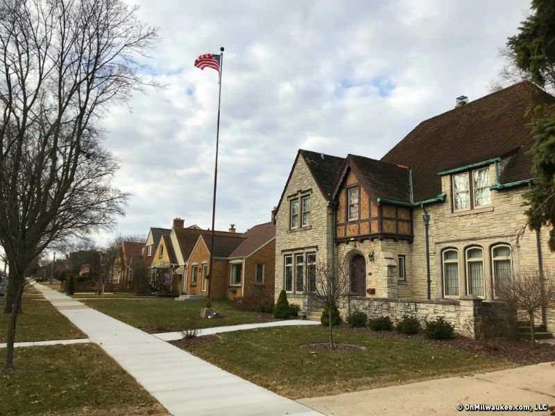 Charming residential street featuring historic homes with gable roofs and stone facades, an American flag on a tall flagpole, and a neatly maintained sidewalk lined with bare trees.