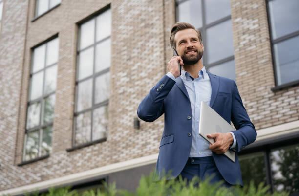 A confident businessman in a blue suit holding a laptop while talking on the phone, standing in front of a modern brick office building."