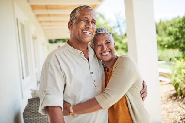 Happy senior couple embracing on the porch of their home, symbolizing comfort and satisfaction in their real estate choice. The image portrays a serene residential setting, perfect for real estate listings targeting retired homeowners.