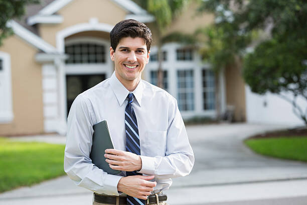 Smiling real estate agent standing outside a modern suburban home, holding a folder and looking professional, ready to assist with property sales.