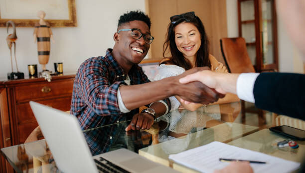 A smiling couple shakes hands with a real estate agent across a table, looking pleased with the agreement they have reached. Documents are visible on the table, indicating the signing of a deal.