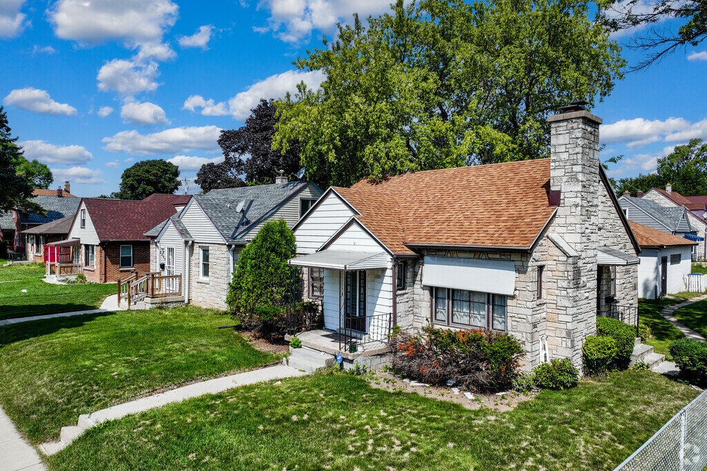 Charming suburban house with a stone facade, gable roof, and a well-maintained front yard, set in a quiet neighborhood with neighboring homes and trees under a bright blue sky.
