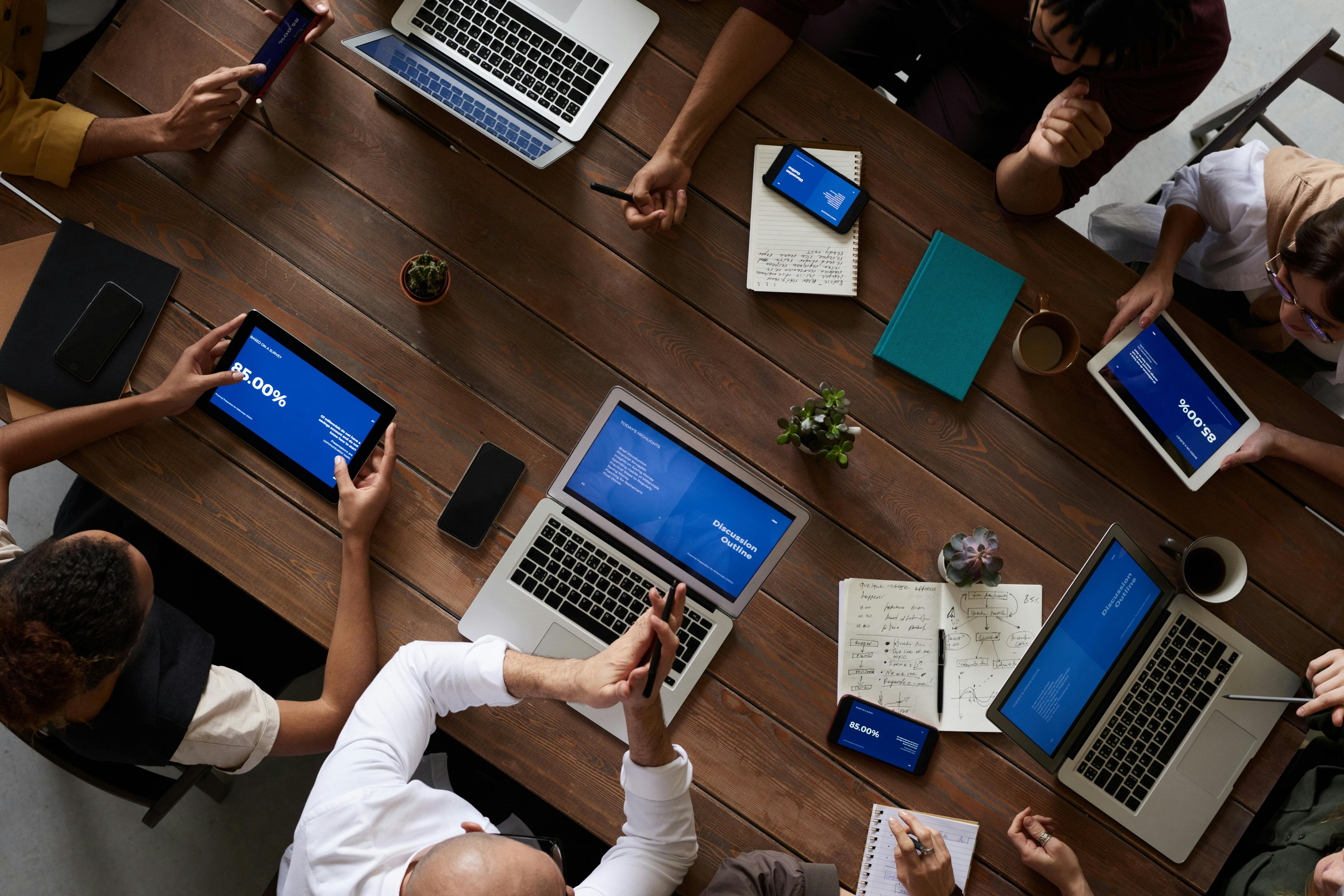 Top view of a team meeting around a wooden table with laptops, tablets, smartphones, notebooks, and coffee cups, displaying data and discussing a project