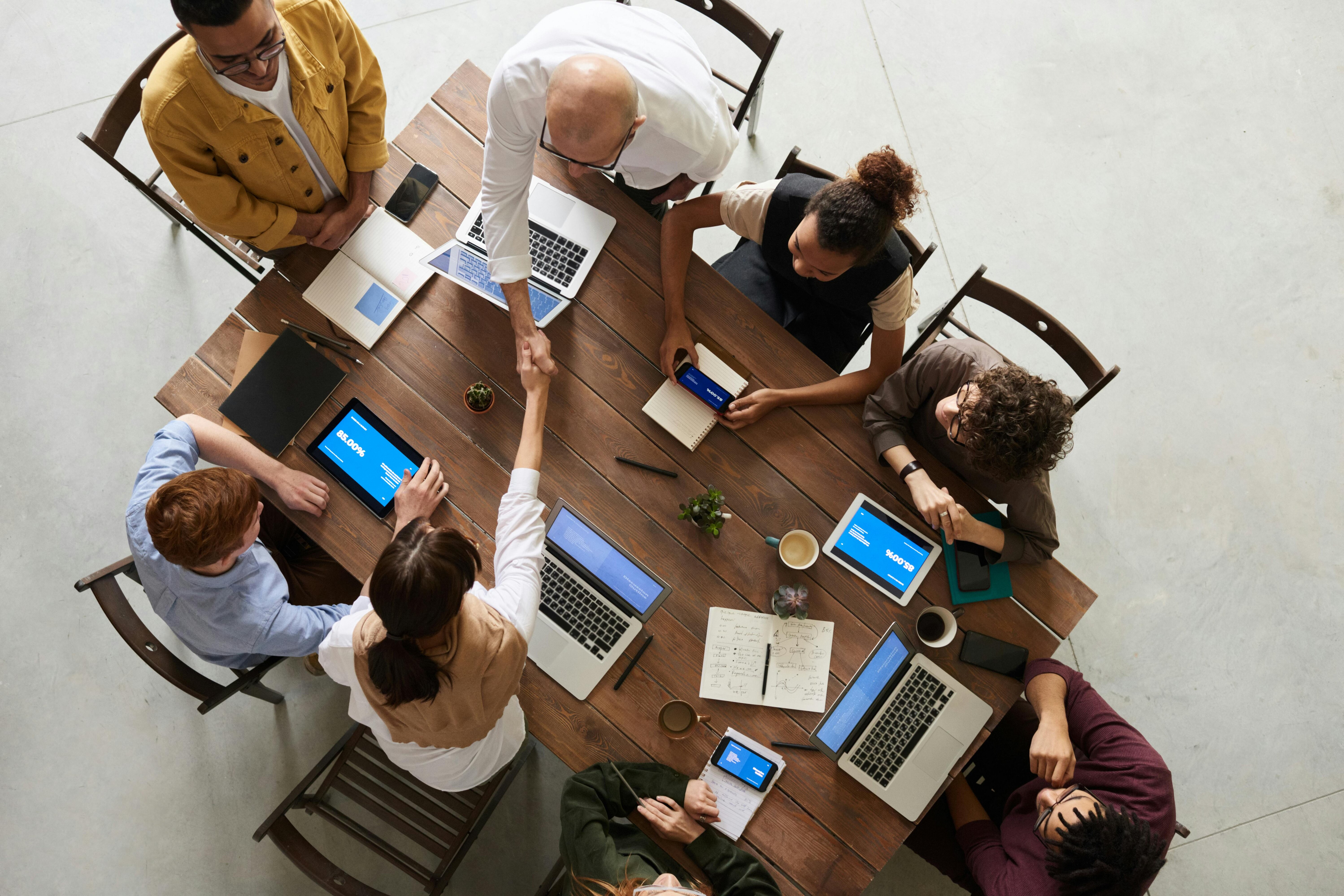 Top view of a diverse team gathered around a wooden table, discussing and working on laptops and tablets. Two members in the center are shaking hands, signifying agreement or collaboration.
