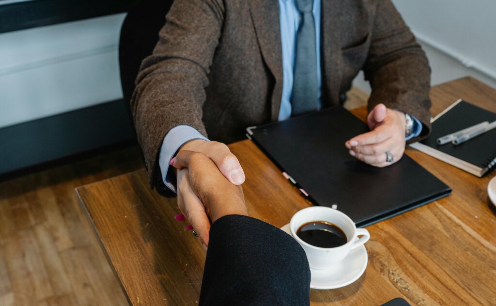 Close-up of two people shaking hands over a wooden table with a cup of coffee, a notebook, and a pen visible, indicating a professional agreement or meeting.