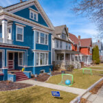 A row of vibrant, historic homes with front porches in a residential neighborhood. The well-maintained lawns feature small soccer goals and a sidewalk runs parallel to the houses, lined with bare trees.