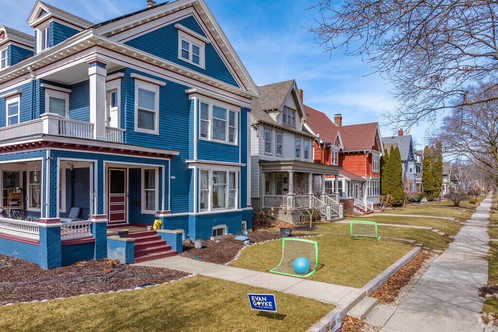 A row of vibrant, historic homes with front porches in a residential neighborhood. The well-maintained lawns feature small soccer goals and a sidewalk runs parallel to the houses, lined with bare trees.