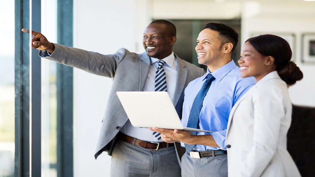 Three business professionals stand together in front of a large window. One man is pointing outwards while the others, a man holding a laptop and a woman, smile and look in the same direction, symbolizing teamwork and future planning.