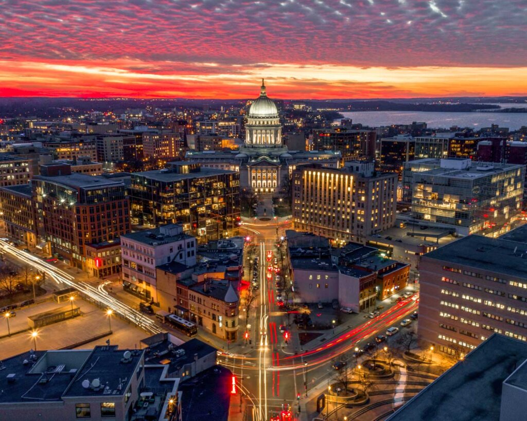 Aerial view of the Wisconsin State Capitol building at sunset, highlighting the bustling city center of Madison with lit streets and a vibrant urban atmosphere.