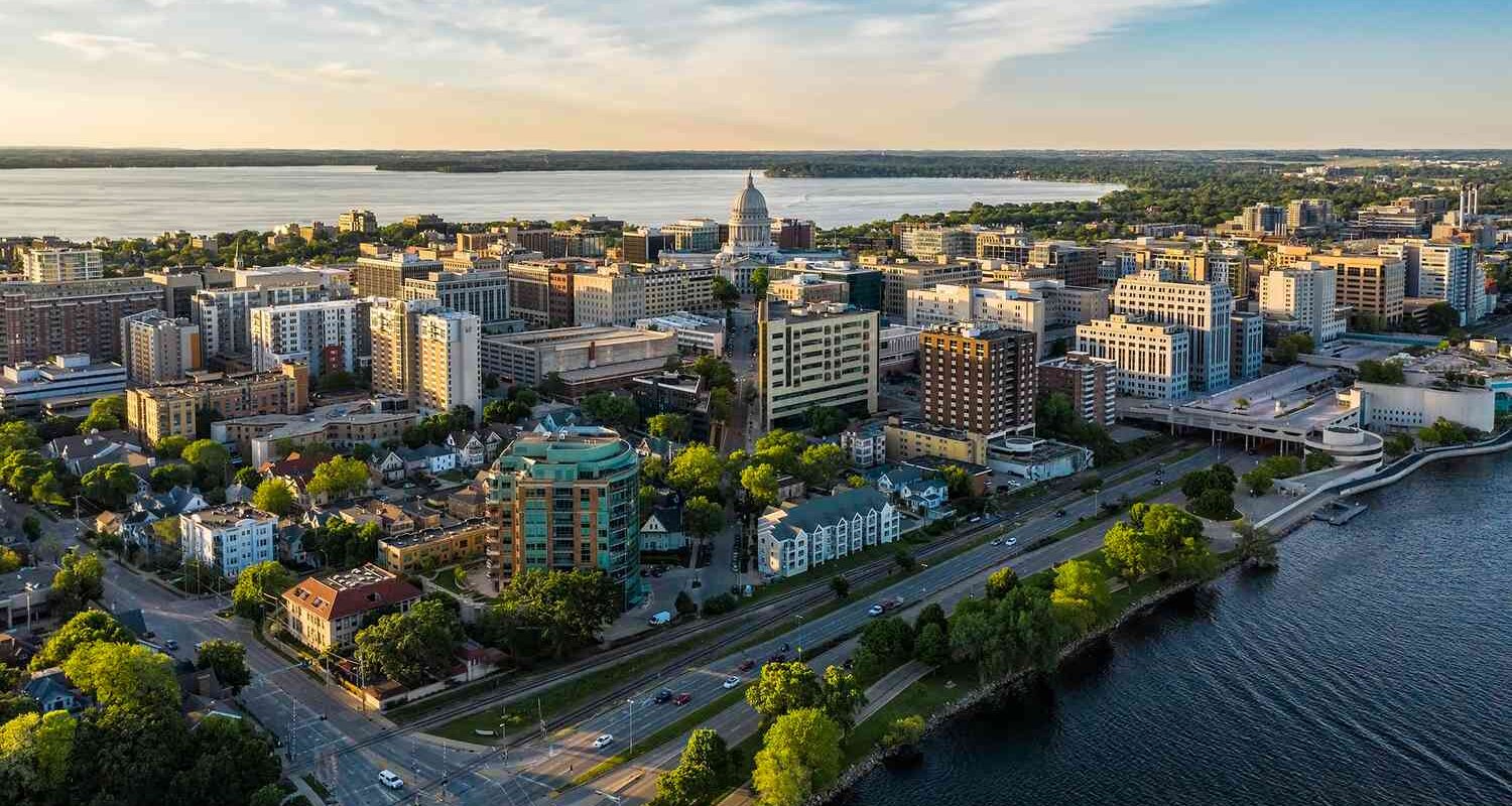 A panoramic aerial view of Madison, Wisconsin, capturing the city’s skyline along the water’s edge, including residential areas and the bustling downtown.