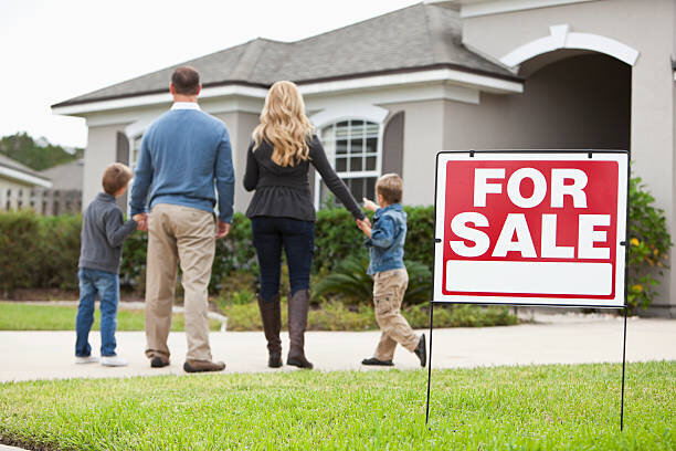A family of four, including two children, stands in front of a house for sale. The parents and children are holding hands while looking at the home, and a "For Sale" sign is visible in the foreground.