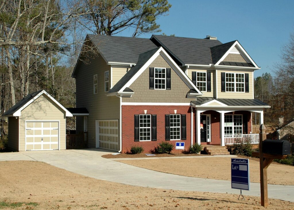 A spacious two-story house with a well-kept driveway, garage, and a "for sale" sign posted in the front yard. The exterior is a mix of brick and siding, and the house has a charming porch.