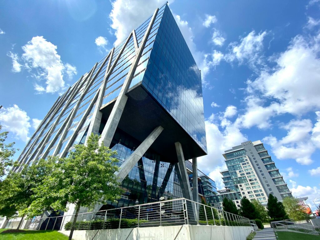Modern glass office building with angled supports, set against a blue sky with clouds.