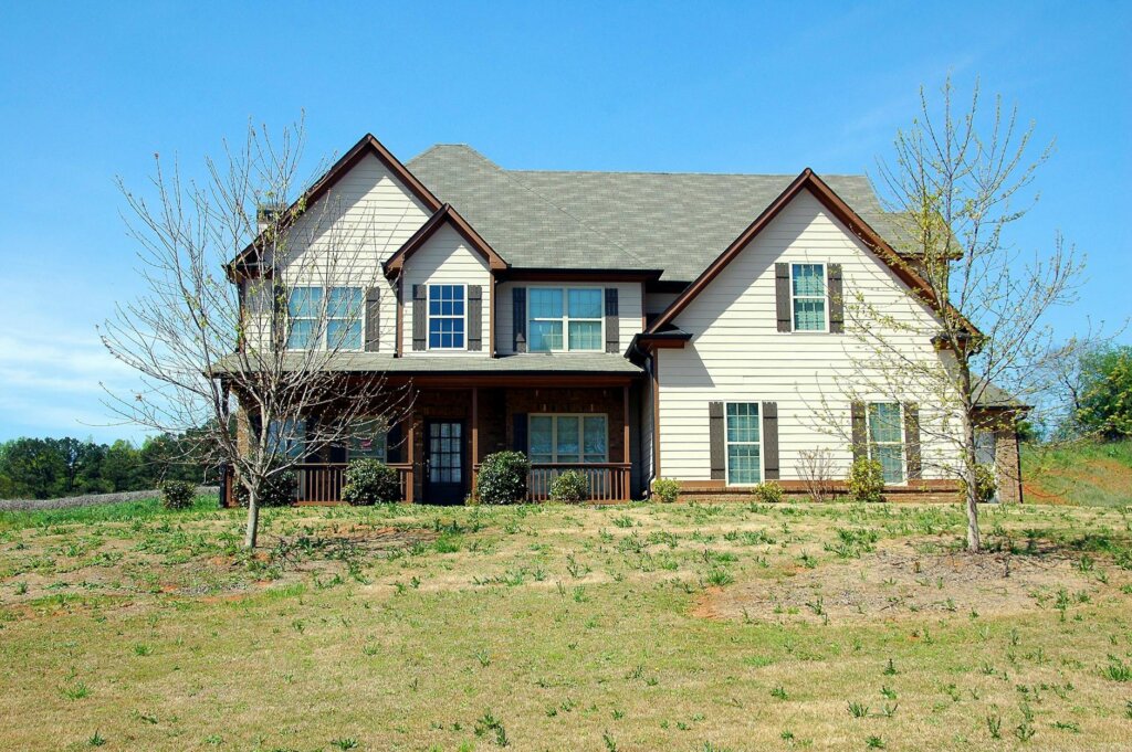 Two-story house with beige siding, dark shutters, and a front porch, set on a grassy lot under a clear blue sky.
