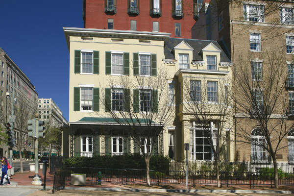 A classic yellow townhouse with green shutters and white trim, set among urban architecture. The house stands out with a traditional design, featuring multiple windows, a front porch, and a fenced-in garden area. The building blends historic charm with the surrounding cityscape.