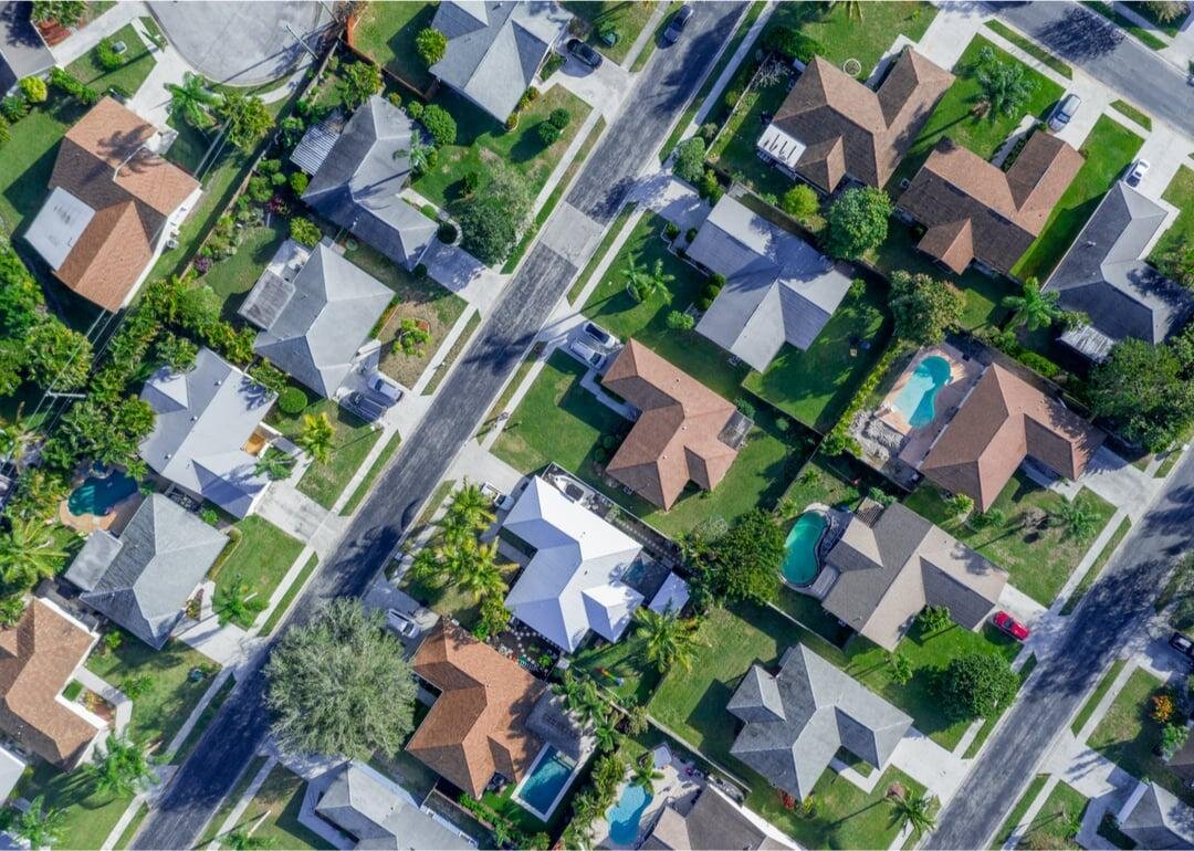Aerial view of a suburban neighborhood with single-family homes, green lawns, and some houses with swimming pools. The street runs through the middle of the image, showing the orderly layout of houses and roads.