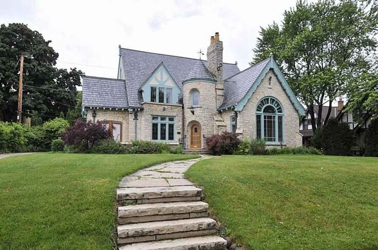 An elegant stone Tudor-style house with distinctive gothic arches and a pointed tower. The house has a slate roof with steep gables and features large arched windows. A stone pathway leads up the well-kept lawn, enhancing the classic, castle-like aesthetic.