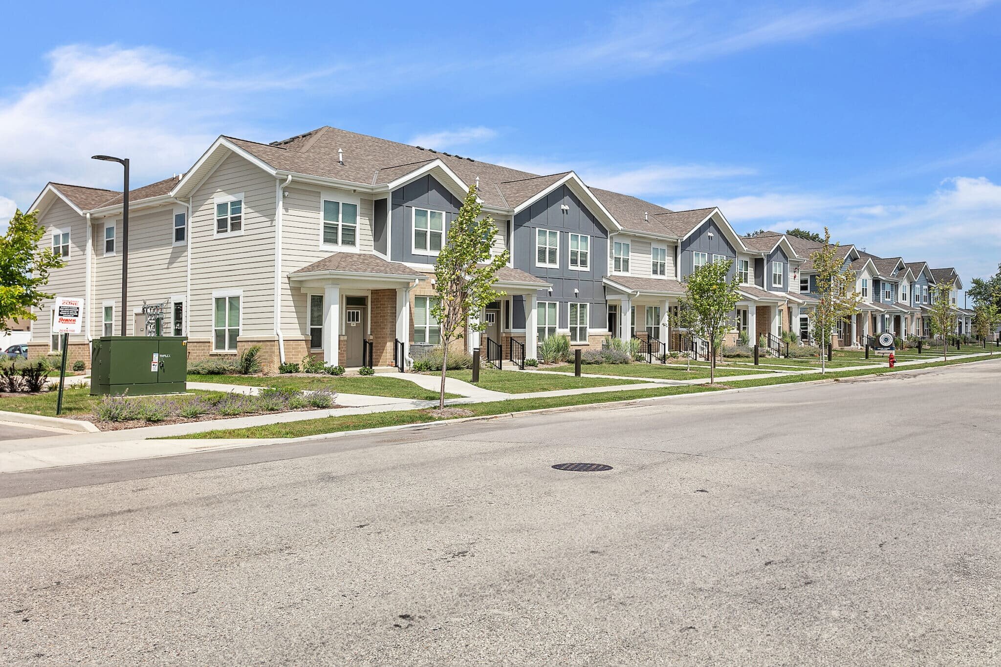 A row of newly constructed modern townhouses with manicured lawns and trees lining the sidewalk. The houses are painted in shades of gray and white, showcasing a clean and welcoming residential neighborhood.