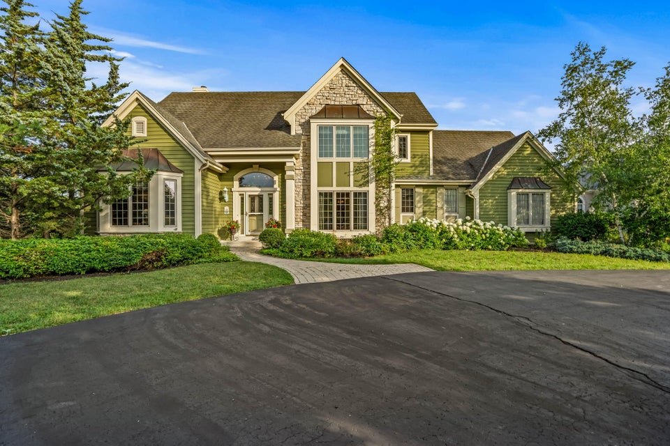  A beautiful green suburban house with stone accents and a tall central window. The front yard has lush landscaping with bushes and trees, and a wide paved driveway leading to the entrance under a clear, sunny sky.