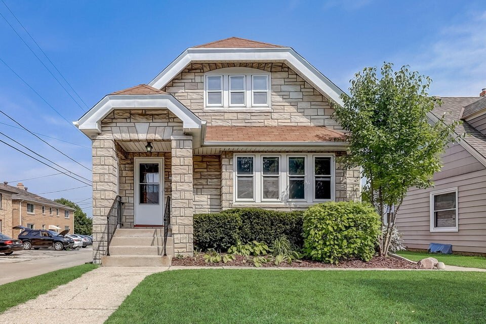 A charming single-story home with a stone facade and steep front steps leading to a white-framed entry door. The house has a gabled roof with brown shingles, large windows, and a neatly landscaped front yard with green bushes and trees, exuding a welcoming suburban feel.