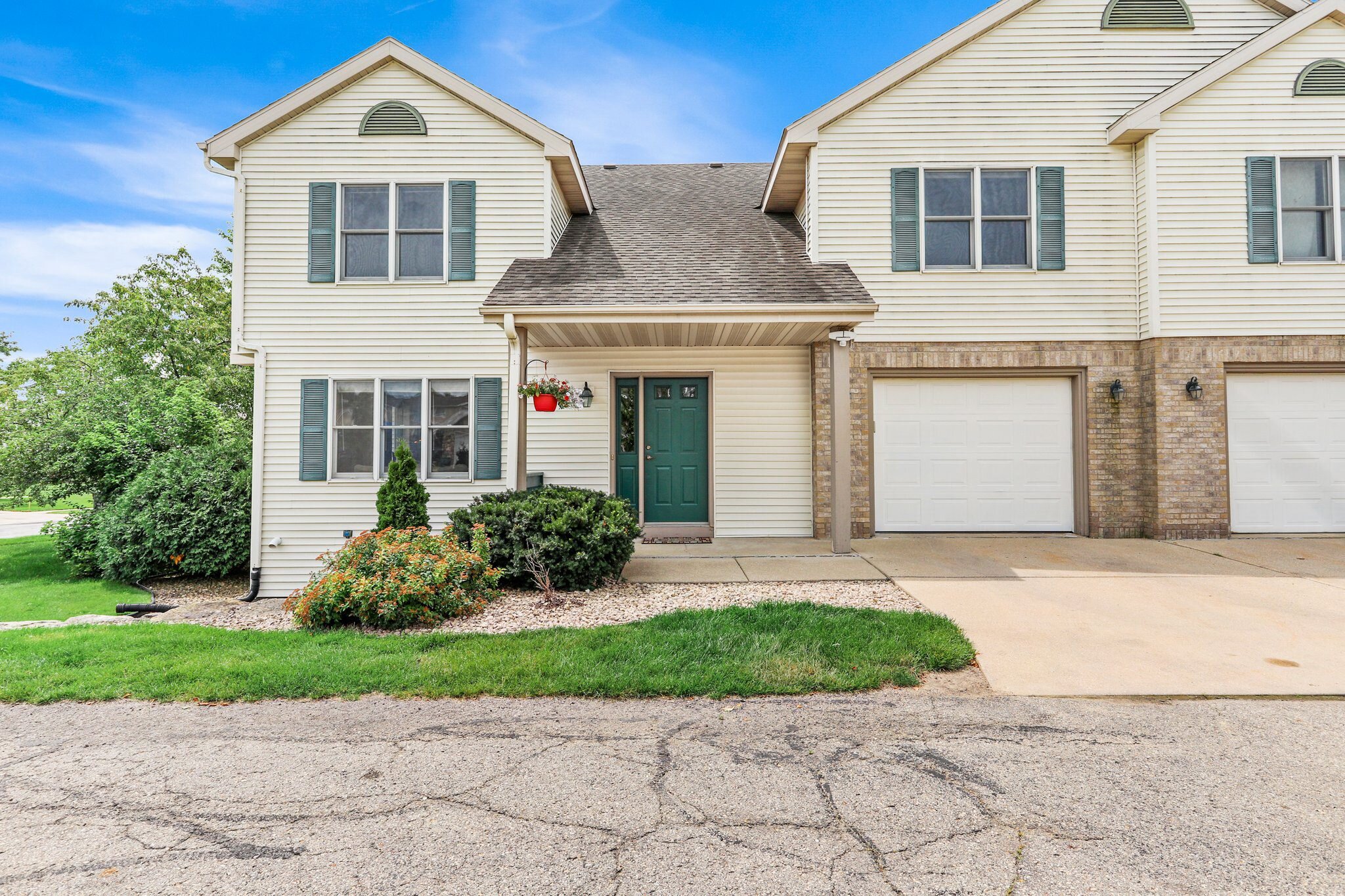 A beige two-story townhouse with a green front door, white garage, and small green shutters on the windows. The front yard has shrubs, and a red hanging flower basket adorns the entryway.