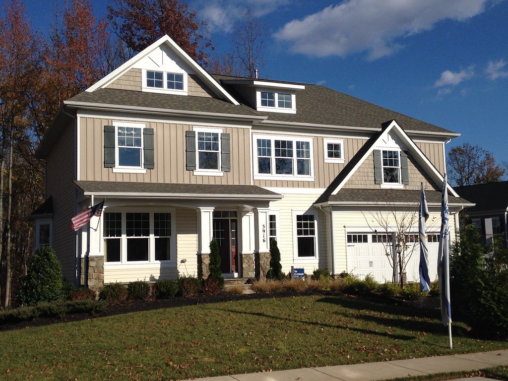 A suburban two-story house with beige and gray siding, a pitched roof, and multiple windows on the front façade. The house has a neatly manicured lawn with small shrubs and an American flag displayed near the entrance, under a clear blue sky.