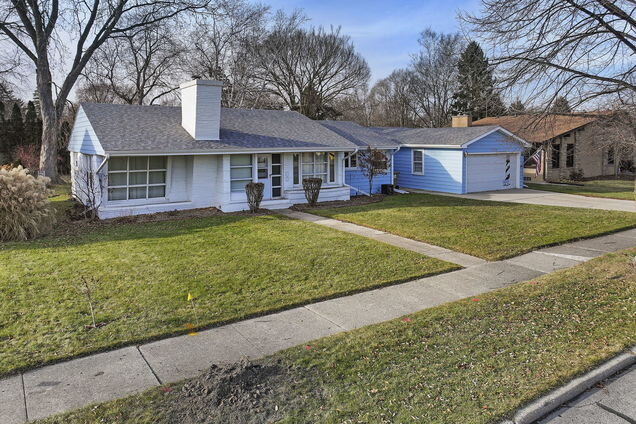 A cozy, single-story blue ranch-style home with a prominent white chimney, surrounded by a spacious green lawn. Sidewalks frame the front yard, leading up to the front door.