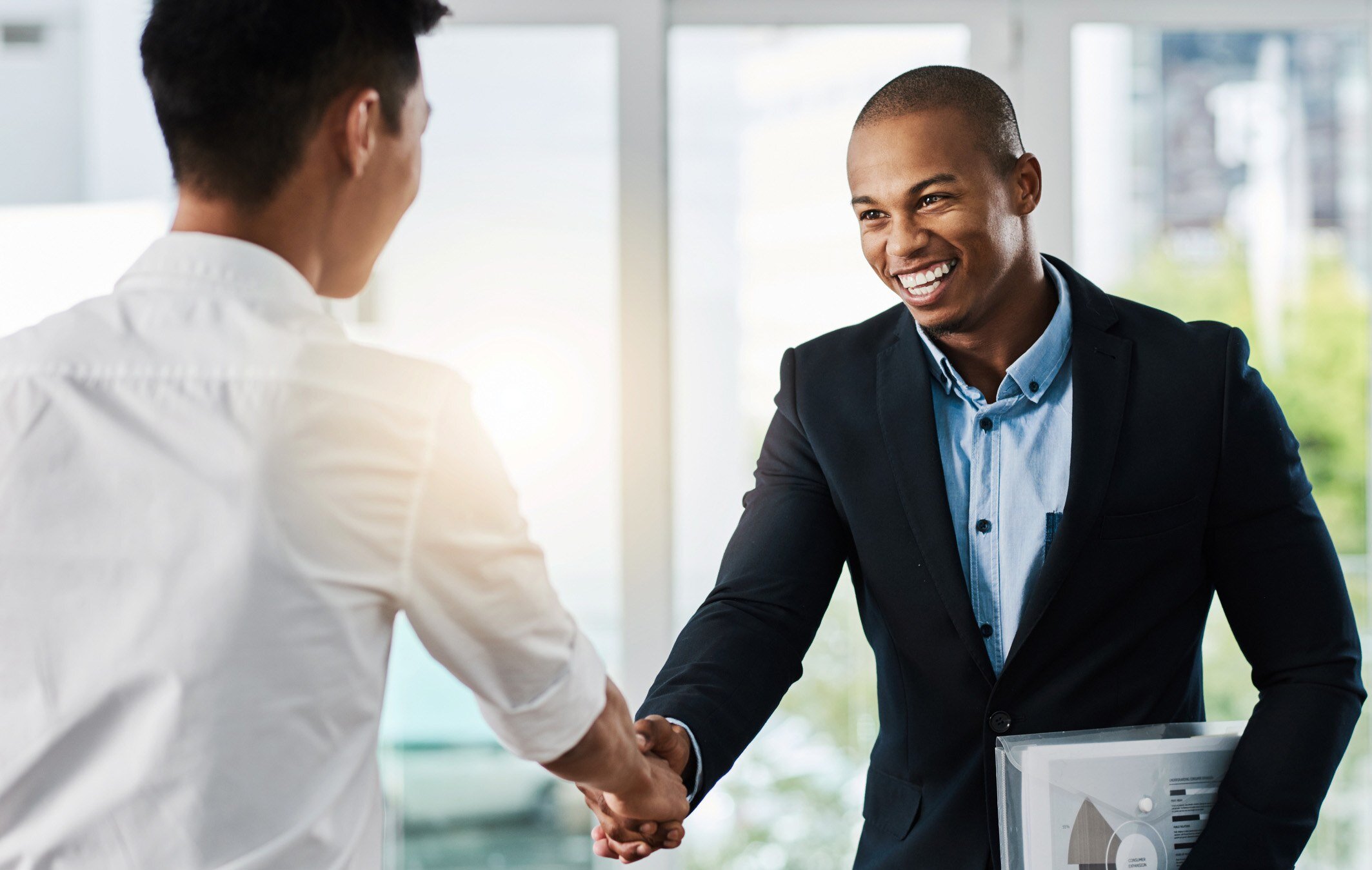A smiling businessman in a dark suit shakes hands with another person, representing a professional agreement or successful partnership.
