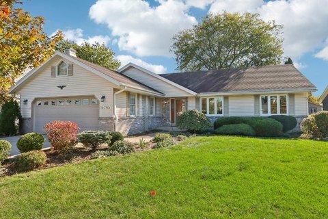 Single-story beige home with an attached garage and a neatly landscaped front yard. The house features a brown shingled roof, large front windows, and decorative shrubs and bushes along the pathway, all under a clear blue sky.
