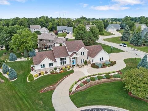 Aerial view of a large, single-story suburban home with a sprawling front lawn and circular driveway. The house has a red roof, white walls, and landscaped garden areas with bushes and trees, situated in a neighborhood of similar homes with green lawns.