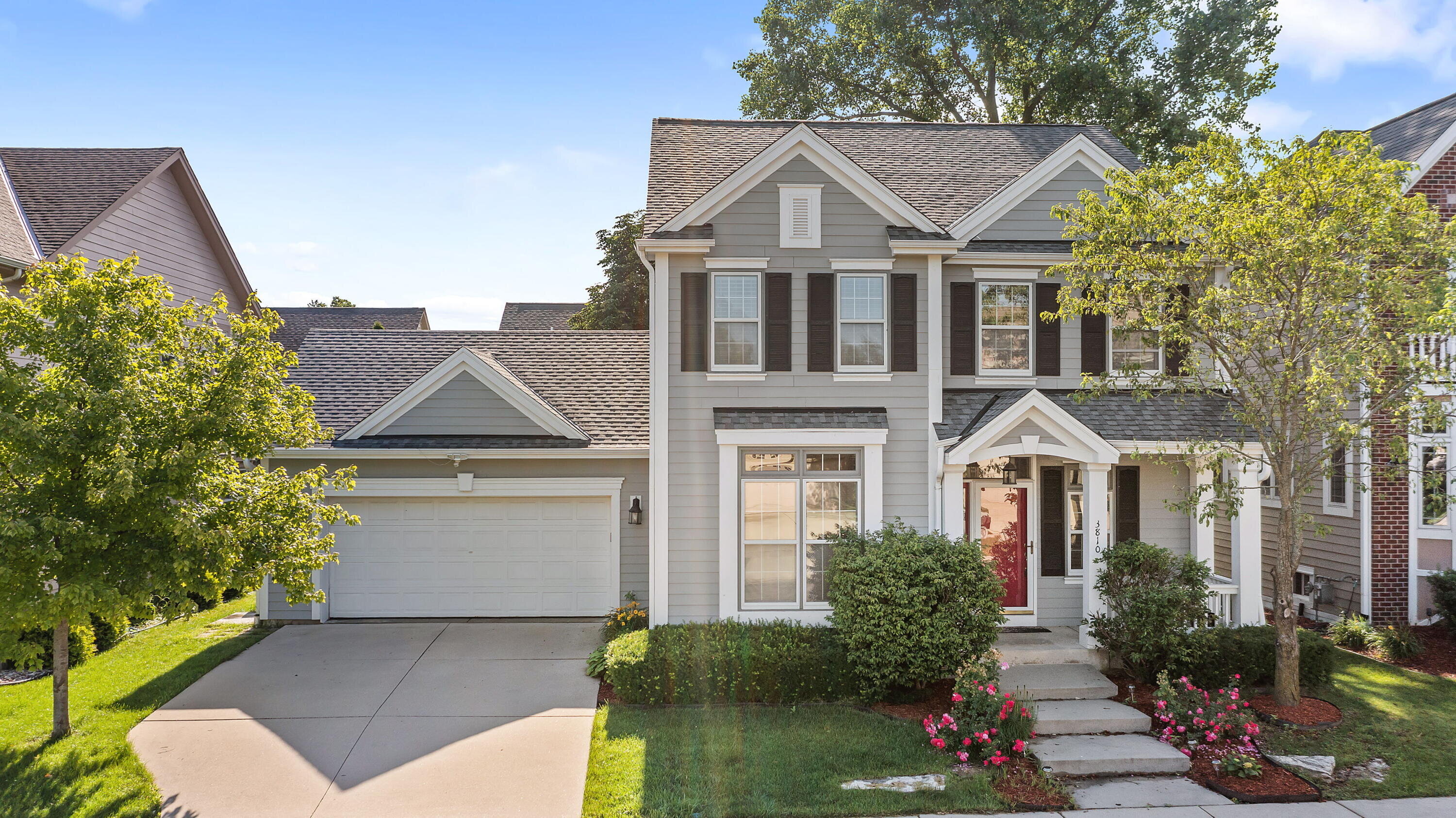 A well-maintained two-story home with a gray facade, white trim, and black shutters, featuring a double garage and a neatly landscaped front yard with vibrant green bushes and blooming flowers under clear blue skies.