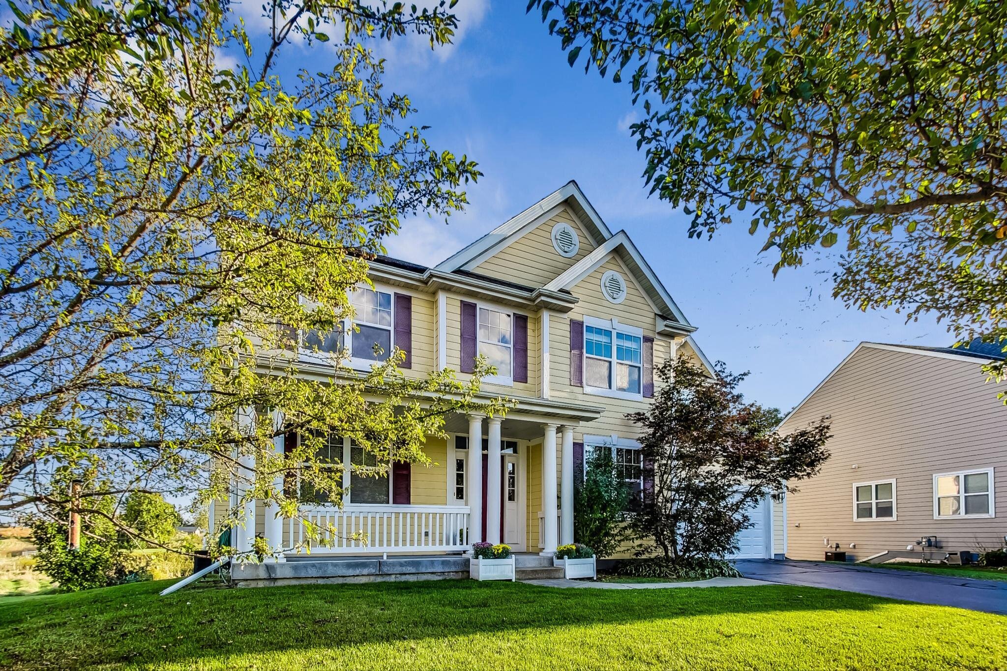 A charming two-story home with light yellow siding, white trim, and red shutters. The house has a spacious porch supported by white columns and is surrounded by lush greenery and trees. A driveway runs along the right side of the house, while the front yard is shaded with tree branches.