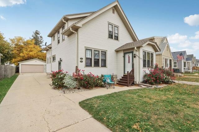 A charming two-story cream-colored home with dark trim and a well-kept driveway leading to a detached garage in the background. The front yard is adorned with bright red flowering bushes, adding a pop of color to the property.