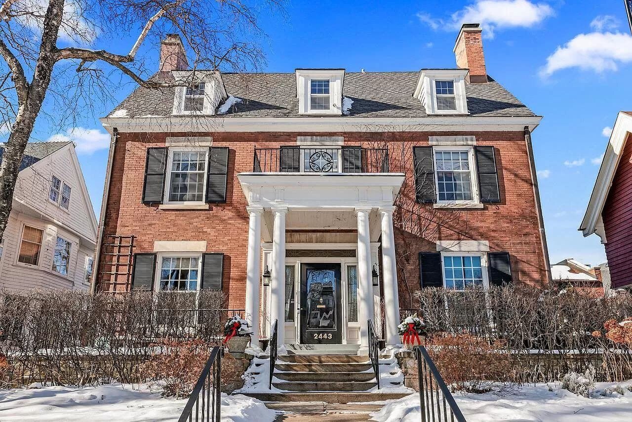 A stately colonial-style brick home with symmetrical architecture, black shutters, and a tall, white-pillared entrance. A staircase with black railings leads up to the front door, decorated with seasonal wreaths. Snow covers the bushes in the foreground, giving a cozy winter feel to the scene.