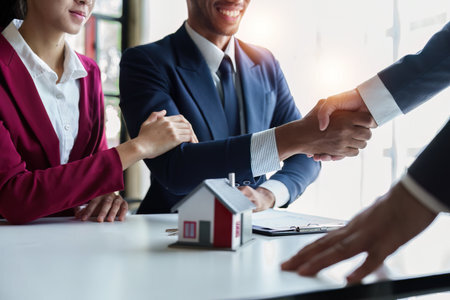 Professional real estate agent shaking hands with clients at a business meeting, symbolizing a successful property agreement. A small model house is placed on the table in front, emphasizing the real estate context.