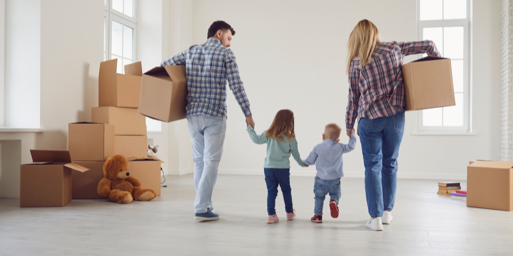 A young family of four, two parents and two children, entering their new home. They are holding hands and walking together through an empty room filled with moving boxes. The scene conveys a fresh start, family togetherness, and the excitement of moving into a new house.