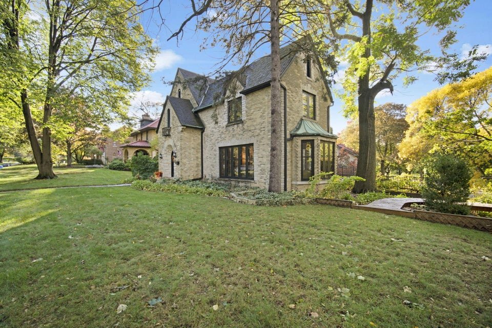 A large, stone Tudor-style house with steep, gabled roofs and elegant dark-framed windows. Surrounded by tall trees and a manicured lawn, this house sits within a lush garden, showcasing a blend of classic architecture and natural beauty.