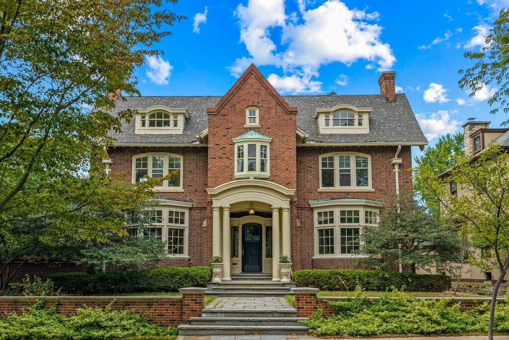 A grand, red-brick, three-story home with a steeply pitched roof and prominent gables. The entrance is adorned with white columns, and large windows provide a glimpse of the spacious interior. A well-manicured garden and small brick pathway lead up to the front entrance, framed by lush green trees