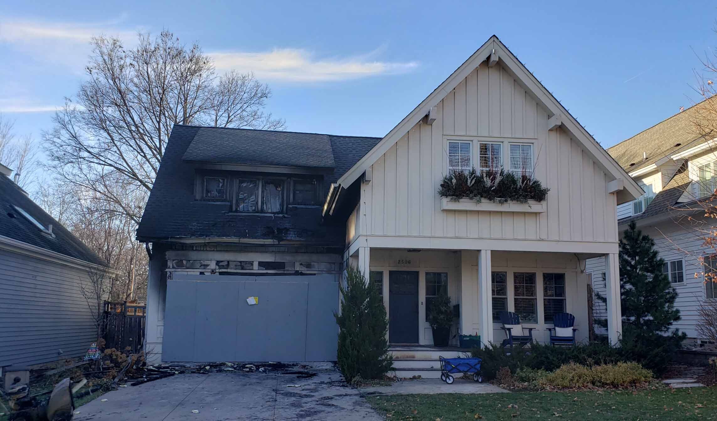  A two-story house with significant fire damage. The left side of the house is charred, with visible structural damage to the roof and garage. The right side appears untouched, contrasting sharply with the burnt section. A stark reminder of the devastating effects of a house fire.