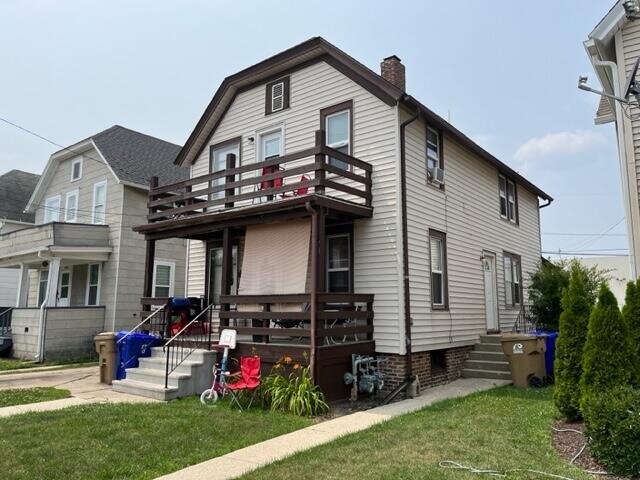 A beige two-story house with a wooden balcony on the second floor, surrounded by a well-maintained lawn. The house is part of a residential neighborhood with sidewalks and a driveway.