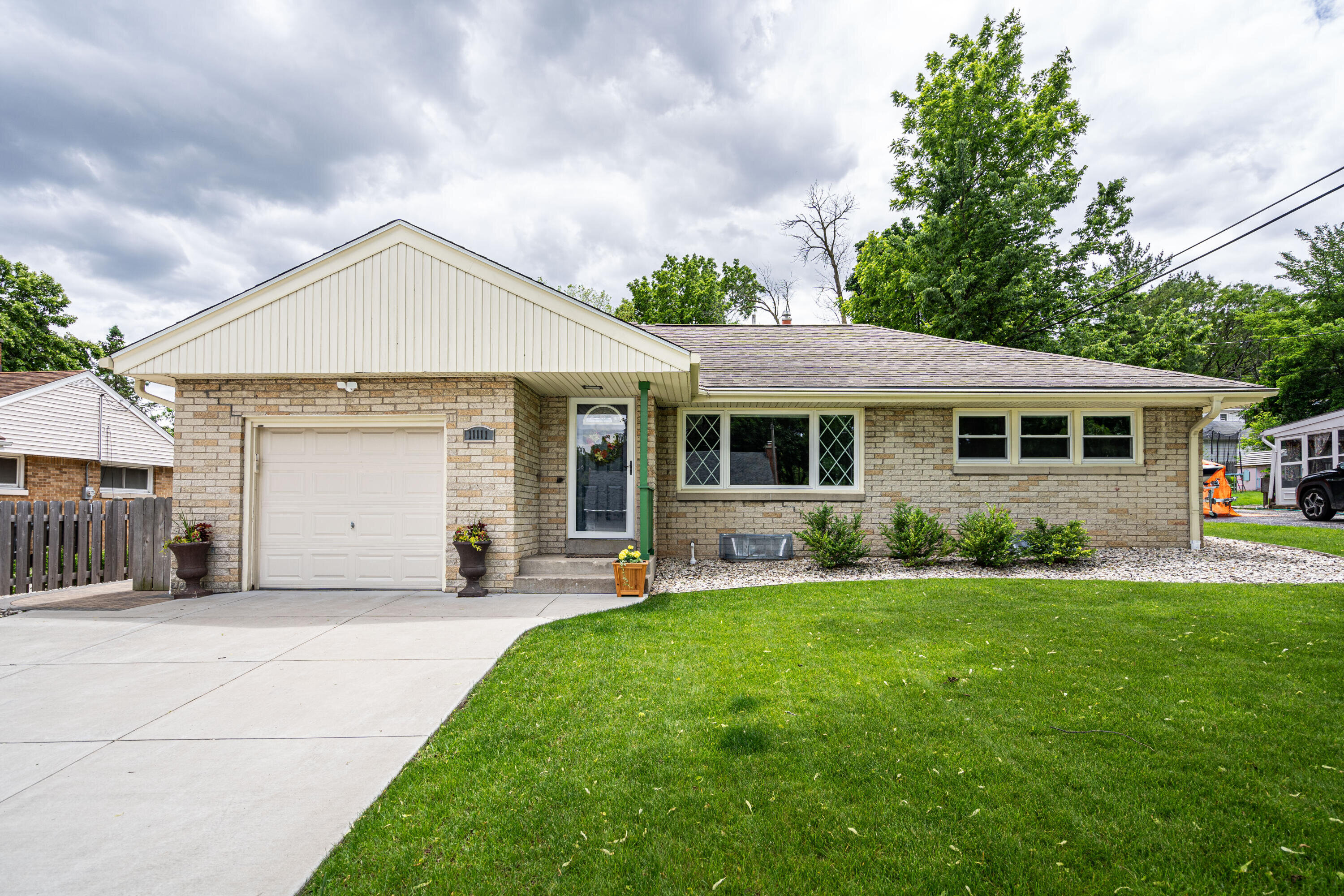 A charming mid-century single-story brick home with a modern front driveway. The house has a neat, welcoming appearance, featuring a light tan brick exterior, a single-car garage, and lush green grass in the front yard.
