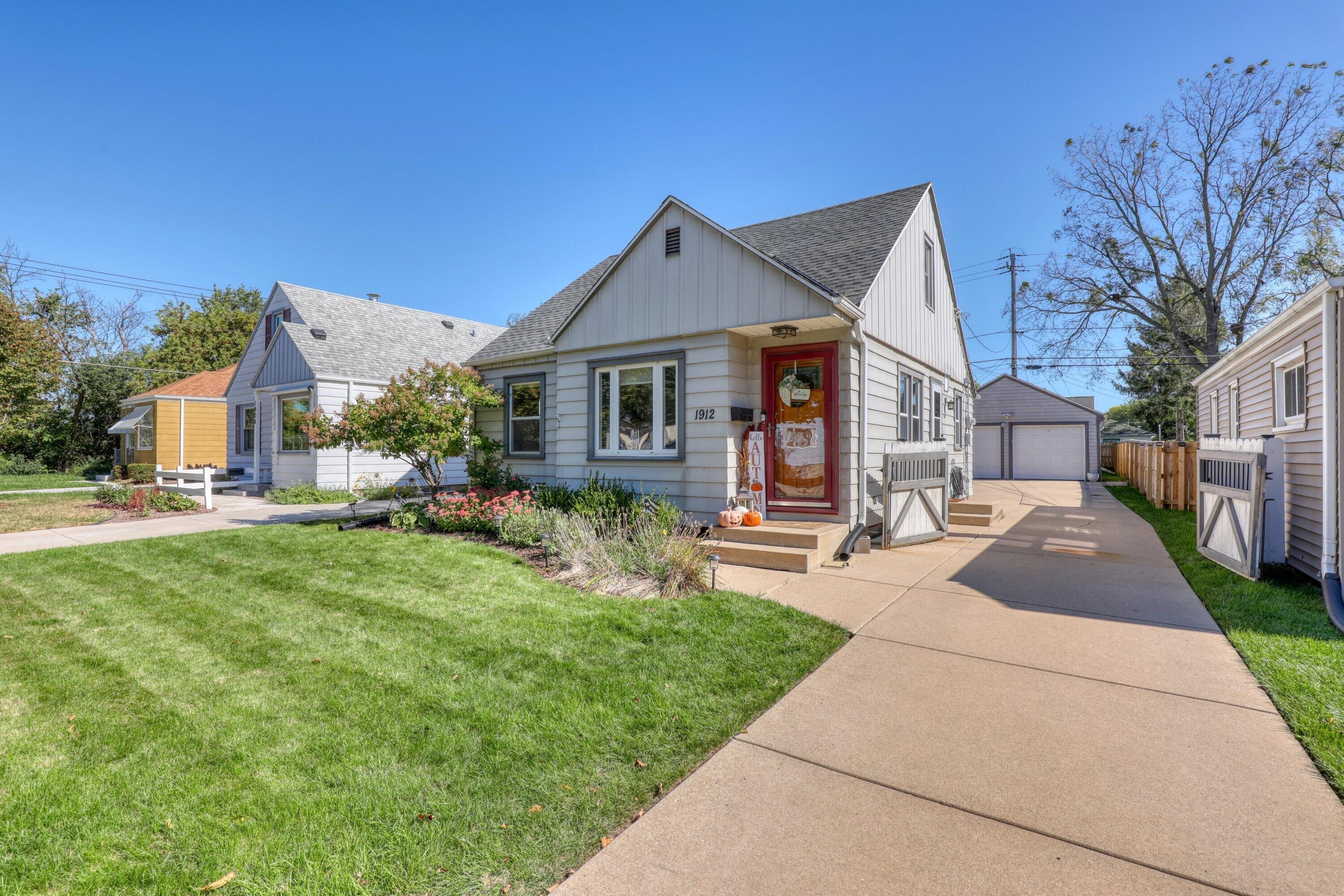 A charming one-story house with gray siding, a bright red door, and seasonal decorations on the porch. A well-maintained lawn and a driveway lead to a detached garage in the background.