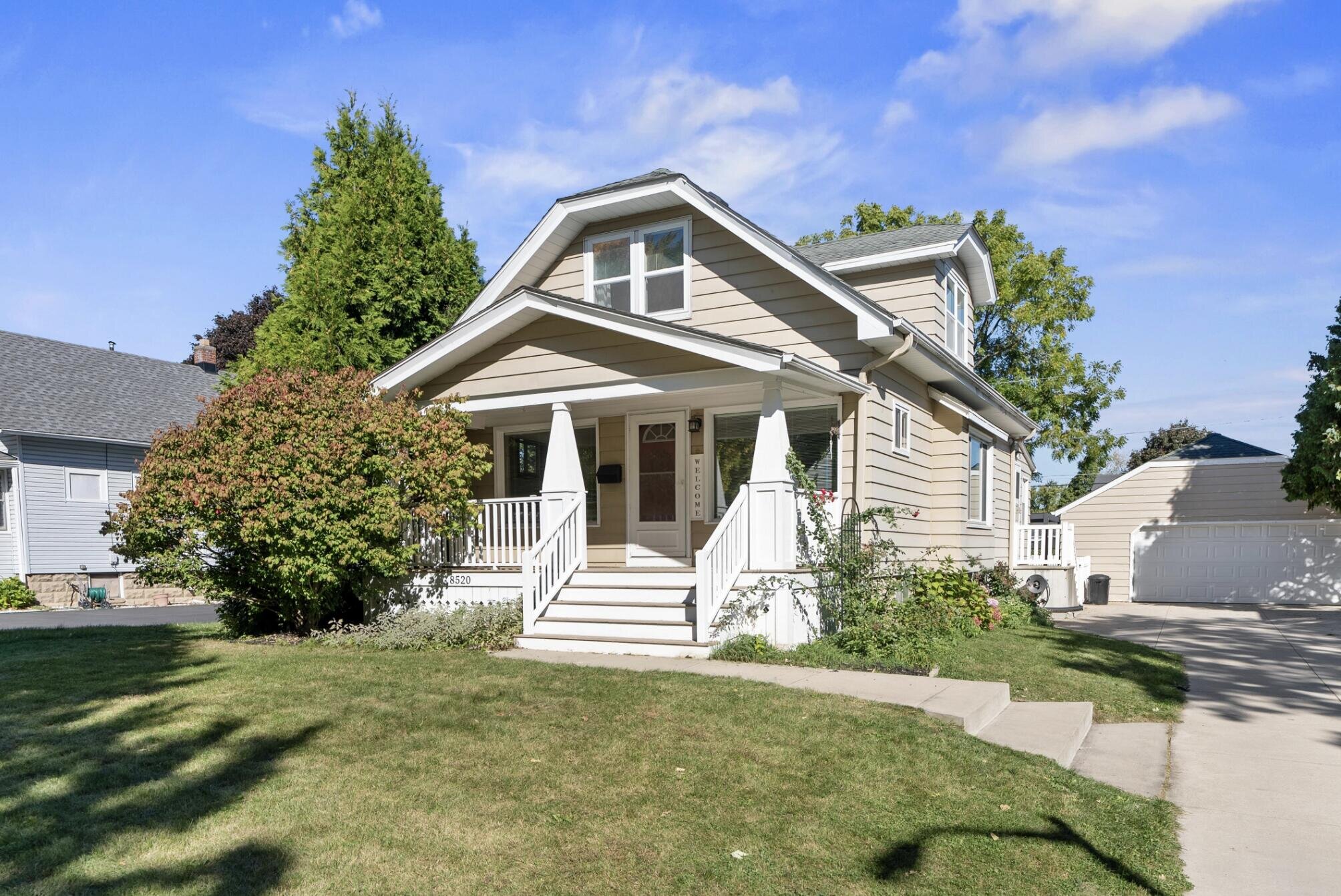 A charming two-story home with beige siding and white trim, featuring a welcoming front porch with steps leading up to it. The driveway on the right leads to a detached garage in the background.