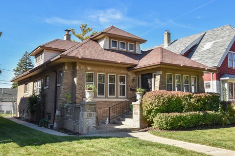 A cozy, single-story brick bungalow with a reddish-brown roof, surrounded by a neatly trimmed lawn and landscaped bushes. The home’s small porch and steps are bordered by stone pillars, adding to its charm in a residential neighborhood setting.